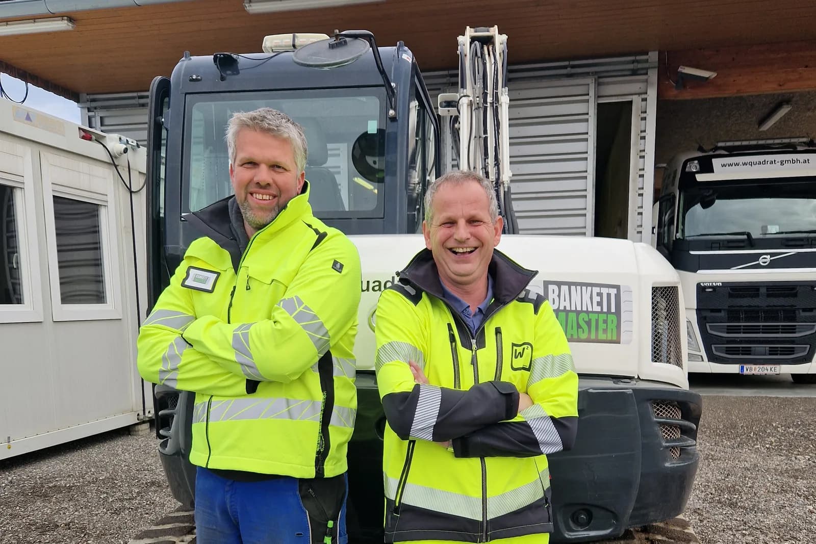 Zwei Mitarbeiter von Bankettmaster in Warnjacken vor einem Bagger am Firmengelände in Niederthalheim